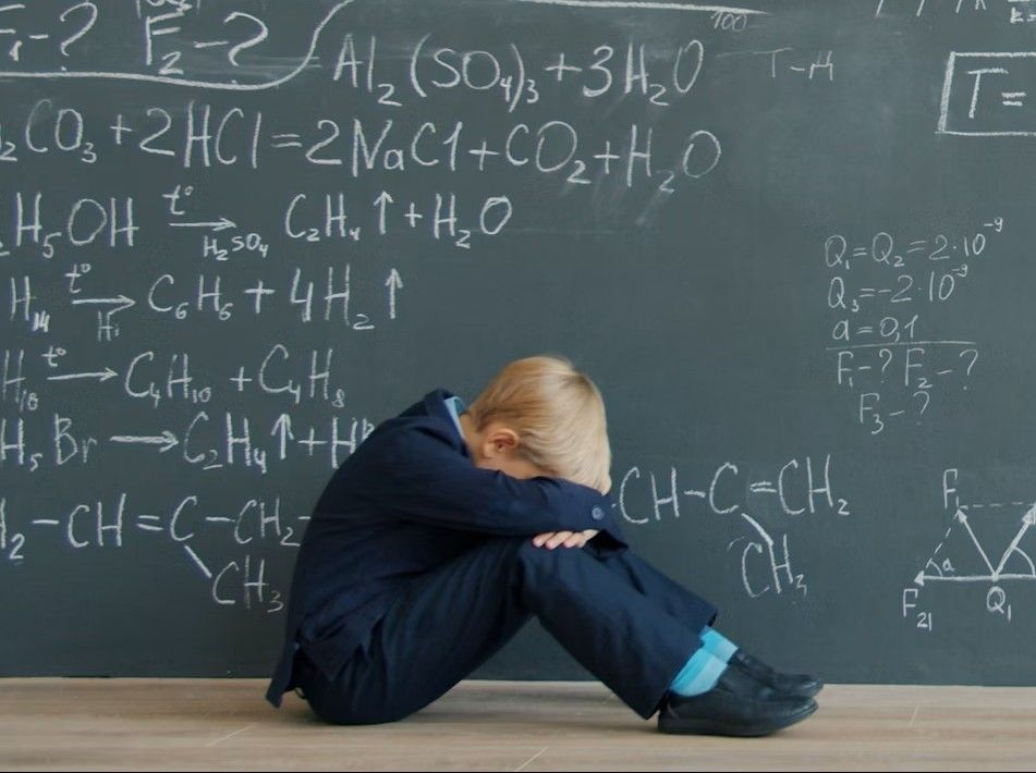 Child sitting on the floor with head down in front of a classroom chalkboard, illustrating school-related anxiety and academic stress in children.