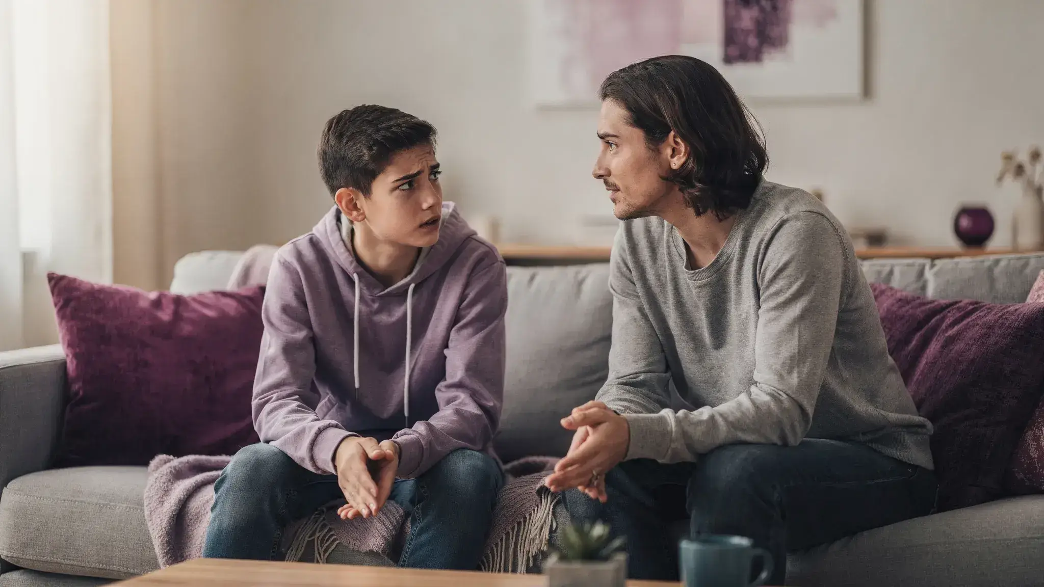 A parent and teenager sitting on a couch in a calm living room, talking face-to-face with open body language; the teen looks frustrated but engaged, and the parent appears supportive and attentive.